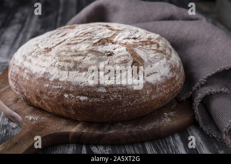 Un pain levain ronde avec le seigle et farine de blé. c'est posé sur une table en bois foncé, avec un fond sombre. Il y a un tour de l'ONU planche à découper Banque D'Images