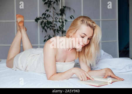 Belle femme blonde lit un livre dans la chambre à coucher au lit Banque D'Images