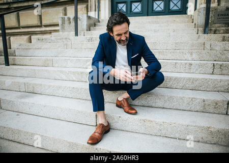 Bearded man wearing blue suit sitting on stairs looking at cell phone Banque D'Images