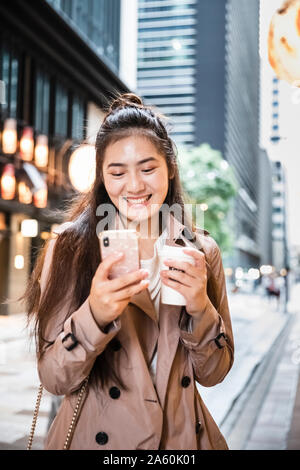 Happy young woman holding boisson à emporter et à l'aide de smartphone dans Ginza, Tokyo, Japon Banque D'Images