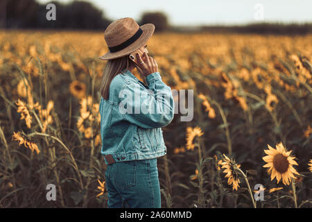 Jeune fille à la veste en jean bleu et un chapeau appelant dans un champ de tournesols dans la soirée Banque D'Images