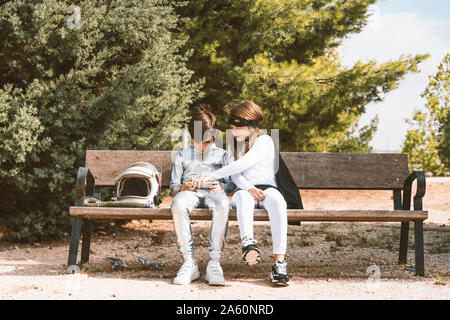 Deux enfants en astronaute et superhero costumes using mobile phone on park bench Banque D'Images