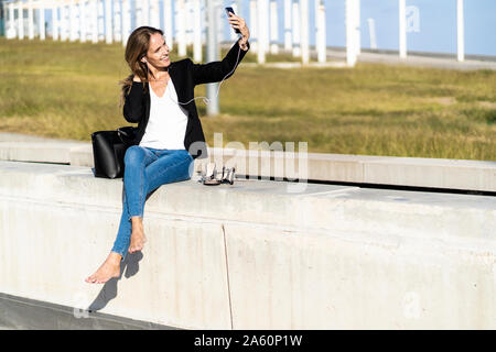 Smiling businesswoman sitting sur un mur avec le smartphone et écouteurs Banque D'Images