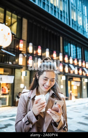 Happy young woman holding boisson à emporter et à l'aide de smartphone dans Ginza, Tokyo, Japon Banque D'Images