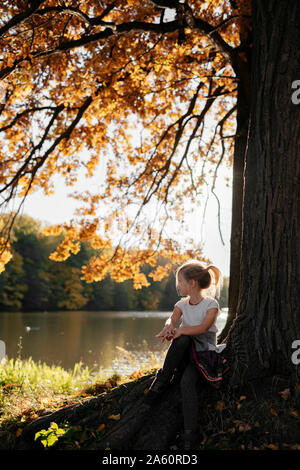 Petite fille assise en vertu de l'arbre d'automne à la recherche à la rivière Banque D'Images