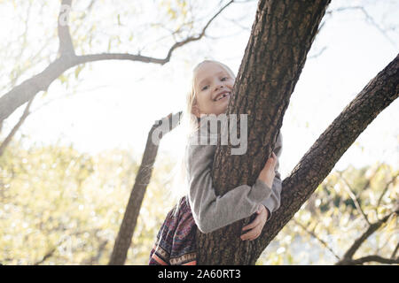 Portrait of smiling little girl climbing sur arbre en automne Banque D'Images