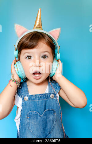 Portrait of cute little girl écouter musique et chant avec écouteurs en forme de licorne sur fond bleu Banque D'Images