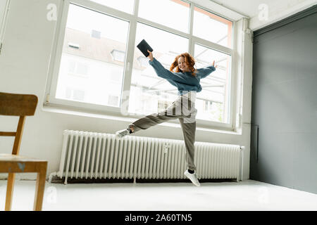 Portrait de femme rousse avec tablette numérique sautant en l'air dans un loft Banque D'Images