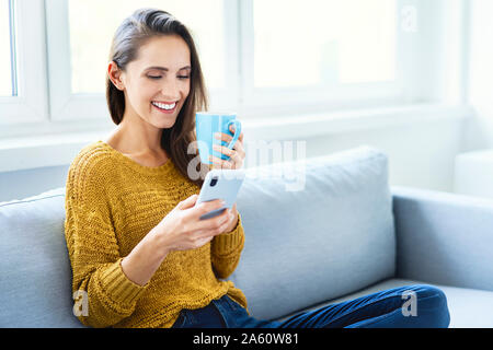 Young woman using smartphone à boire du café et sitting on sofa Banque D'Images