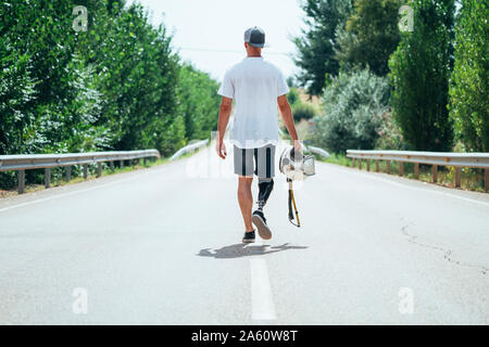 Jeune homme avec prothèse de jambe marchant le long de la route avec un casque Banque D'Images