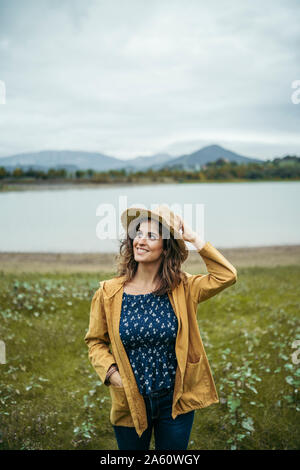 Jeune femme aux cheveux bouclés portant un manteau jaune et bleu t-shirt tenant un chapeau dans sa tête avec un lac à l'arrière-plan Banque D'Images