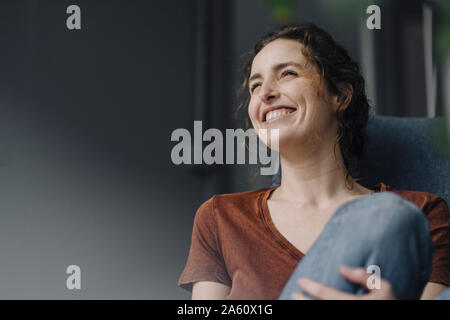 Portrait of laughing young woman sitting on lounge chair at home Banque D'Images