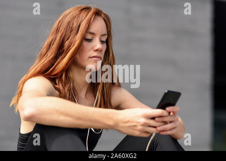 Jeune femme sportive avec des écouteurs ayant une pause en utilisant smartphone outdoors Banque D'Images