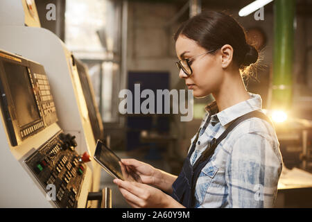Opératrice en lunettes, debout près de la machine et le contrôle du travail d'elle with digital tablet in factory Banque D'Images