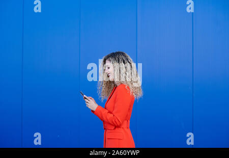 Profile of young woman wearing red dress devant fond bleu looking at cell phone Banque D'Images