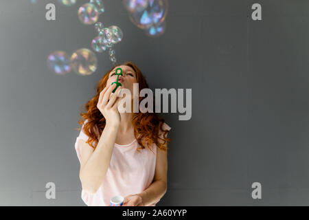 Redheaded woman leaning against wall gris soufflant des bulles de savon Banque D'Images