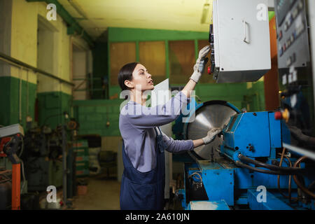 Jeune travailleuse en salopette debout et appuyer sur des boutons sur la machine pendant qu'elle travaille dans l'usine Banque D'Images