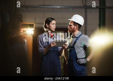 Jeune travailleuse en salopette holding digital tablet et parler à son collègue pendant qu'ils travaillent dans l'usine Banque D'Images