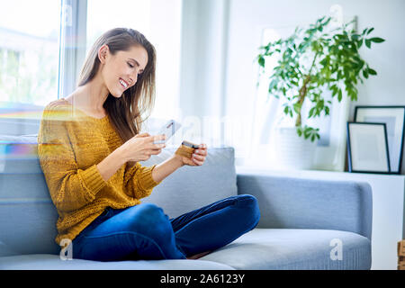 Belle femme heureuse de payer en ligne avec carte de crédit et smartphone while sitting on sofa Banque D'Images