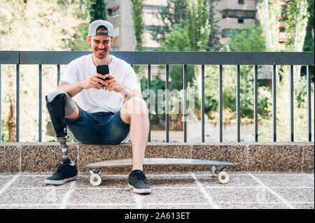 Smiling young man avec prothèse de jambe et skateboard using smartphone Banque D'Images