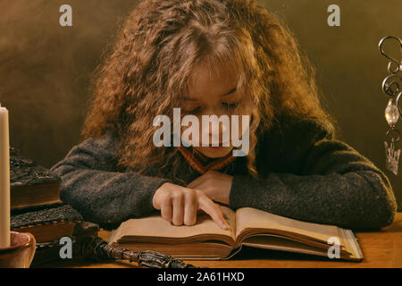 Portrait of a cute enchantress enfant avec de magnifiques longs cheveux bruns vêtu d'un pull bleu marine, chemise blanche et cravate rouge. Elle est en train de lire un livre wi Banque D'Images