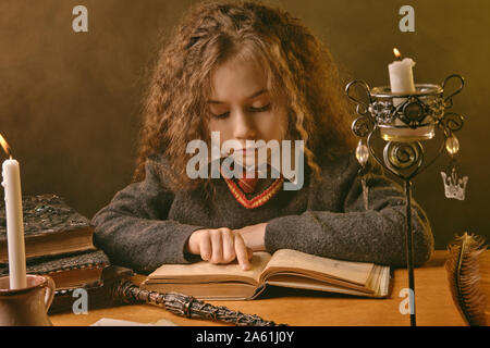 Portrait of a cute kid sorcière avec de magnifiques longs cheveux bruns vêtu d'un pull bleu marine, chemise blanche et cravate rouge. Elle est en train de lire un livre avec s Banque D'Images