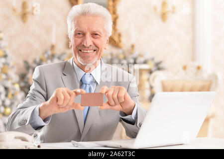 Portrait of happy senior man sitting at table with laptop and holding blank business card Banque D'Images