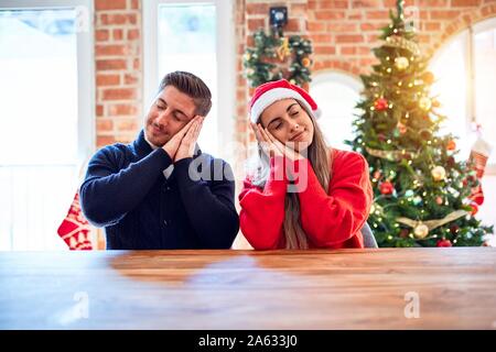 Jeune couple wearing santa claus hat assis sur une chaise et une table autour de l'arbre de Noël à la maison fatigué dormir rêver et posant avec les mains ensemble w Banque D'Images