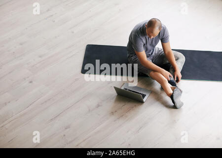 Jeune homme handicapé avec une prothèse à la jambe assis sur les tapis d'exercice et à l'aide d'ordinateur portable avant la formation Banque D'Images