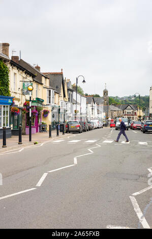 L'homme marchant sur un passage pour piétons, avec des voitures en stationnement le long de la rue principale de Bridgend, Glamorgan, Pays de Galles, Royaume-Uni Banque D'Images