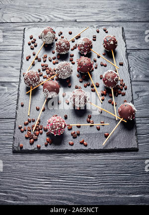 Cake pops au chocolat maison saupoudré de bonbons et de noix de coco broyée sur sprinkles une pierre noire plateau avec café, vue verticale Banque D'Images