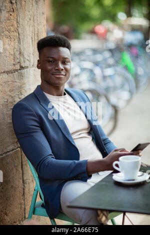 Businessman s'asseoir à une table d'extérieur dans une ville tout en tapant sur son téléphone portable et regardant la caméra. Banque D'Images
