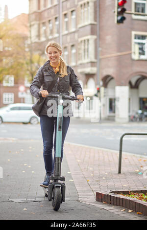 Young happy blonde woman riding scooter électrique dans la ville, souriant à la caméra, à l'automne Banque D'Images