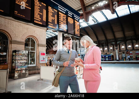 Un businessman and businesswoman Standing together dans une gare. La femme est montrant l'homme quelque chose sur son téléphone mobile. Banque D'Images