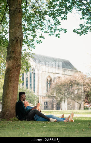 Un homme et une femme assise dans un parc contre un arbre. La femme est assise entre les jambes tout en lui montrant quelque chose sur son téléphone mobile. L Banque D'Images