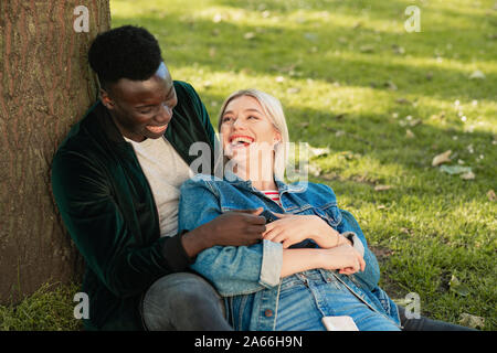 Un homme et une femme assise dans un parc contre un arbre. L'homme tient à elle comme elle est assise entre ses jambes. Ils sont à la recherche à l'autre tout en laughin Banque D'Images