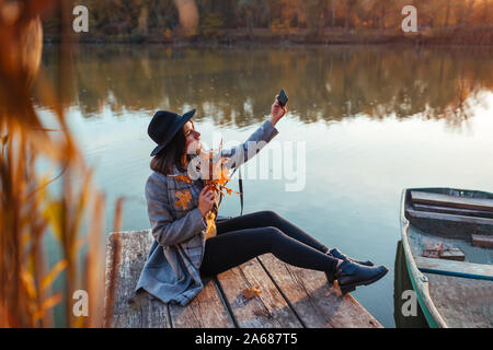 Saison d'automne. Prendre Femme avec selfies rameaux avec feuilles à l'aide du smartphone sur le quai de la rivière. Réseau social des toxicomanies Banque D'Images