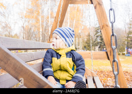 Tout-petit enfant regarde autour de lui avec intérêt swinging sur balançoire en bois par beau jour d'automne dans la région de City Park. Banque D'Images