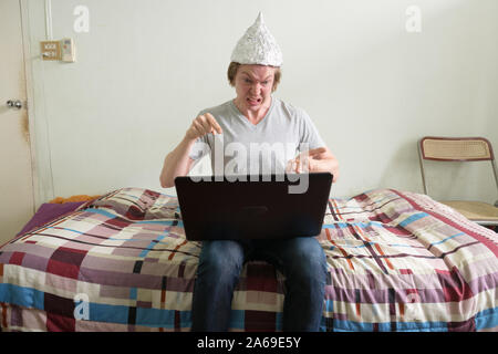 Jeune homme a souligné avec du papier d'aluminium hat en utilisant ordinateur portable et à la colère dans la chambre à coucher Banque D'Images