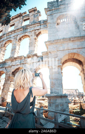 Jeune femme prend des photos touristiques de Pula Arena, la Croatie avec son smartphone sur belle journée ensoleillée. Tiré de l'arrière. Destinations de voyage européen Banque D'Images