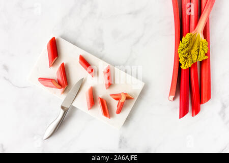 Couper les jeunes la rhubarbe forcée sur pièces en planche à découper avec couteau et tiges de rhubarbe avec la feuille sur fond de marbre blanc avec l'exemplaire de l'espace. Vue d'en haut. Banque D'Images
