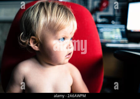 Une jolie petite fille aux cheveux blonds et aux yeux bleus lumineux assis sur une chaise de bureau rouge, environ 18 mois Banque D'Images