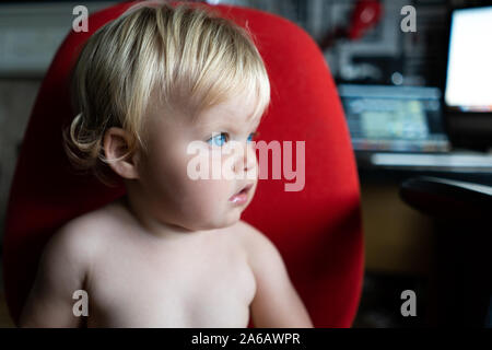 Une jolie petite fille aux cheveux blonds et aux yeux bleus lumineux assis sur une chaise de bureau rouge, environ 18 mois Banque D'Images