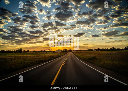 Un pays route pavée menant à l'Horizon comme les rayons du soleil beam hors des nuages au lever ou au coucher du soleil Banque D'Images