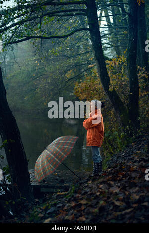 Une femme dans un manteau de pluie orange regarde le lac et la beauté de la nature. Parapluie, l'automne. Banque D'Images