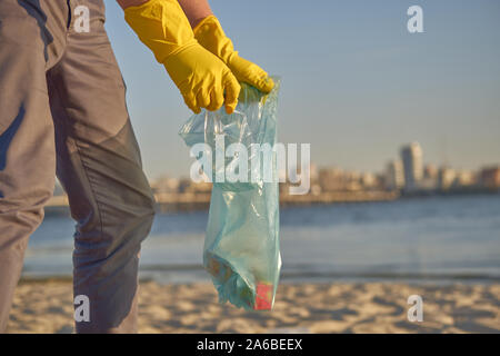 Responsable bénévole dans des gants de caoutchouc jaune marche avec sac à déchets le long d'une rive de la rivière sale et le nettoyage de la corbeille. Les gens et l'écologie. Banque D'Images