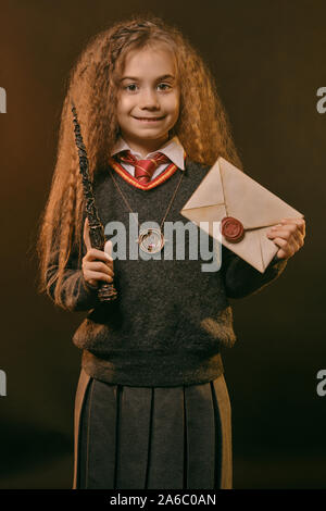 Portrait of a cute little charmer avec de magnifiques longs cheveux bruns vêtu d'un pull bleu marine et gris jupe. Elle sourit, tenant une baguette magique Banque D'Images