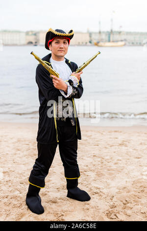 Young man in a black pirate costume bras croisés avec les armes-jouets en face de lui sur une plage de sable. La verticale. Banque D'Images