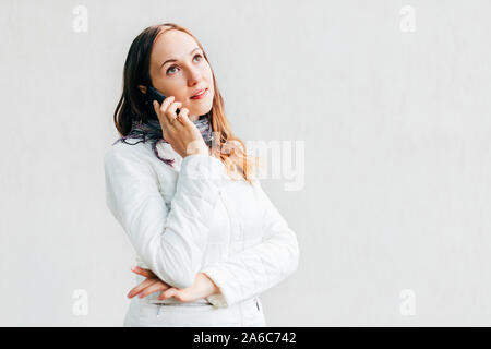 Portrait Portrait de jeune femme en veste blanche à l'écart pensivement et talking on mobile phone sur fond monochrome solide mur. Banque D'Images