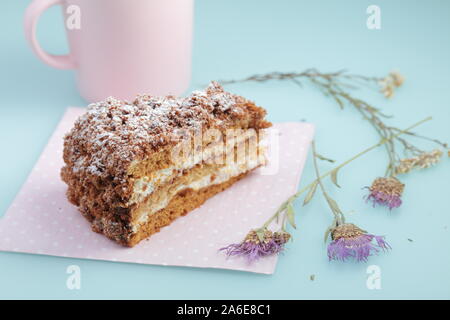 Tranche de gâteau russe medovik avec glaçage sur fond bleu d'œuf de canard avec fleurs séchées et tasse de lait rose. Couleurs pastel et espace de copie. Banque D'Images